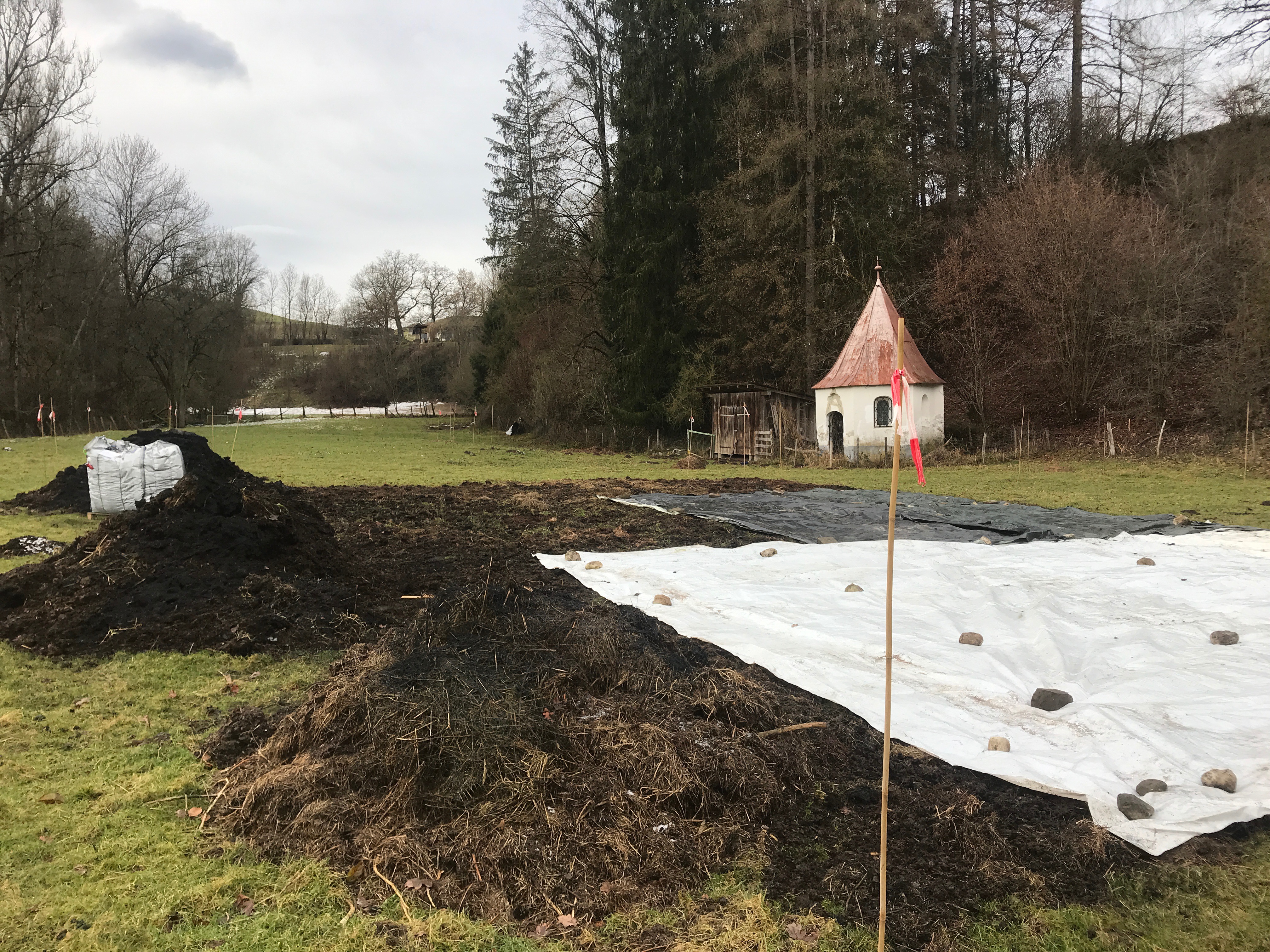 Grünland mit aufgebrachtem Kompost und Silofolie und Kapelle am Hangfuß im Hintergrund